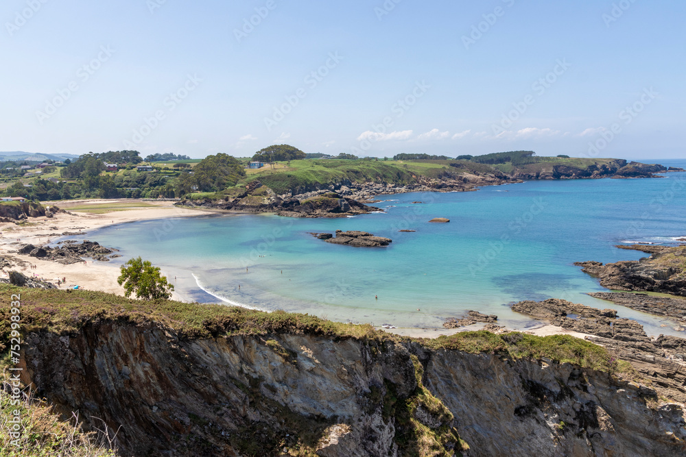 a serene beach with turquoise waters, surrounded by rocky terrain and lush greenery under a clear sky. A few people are visible in the distance