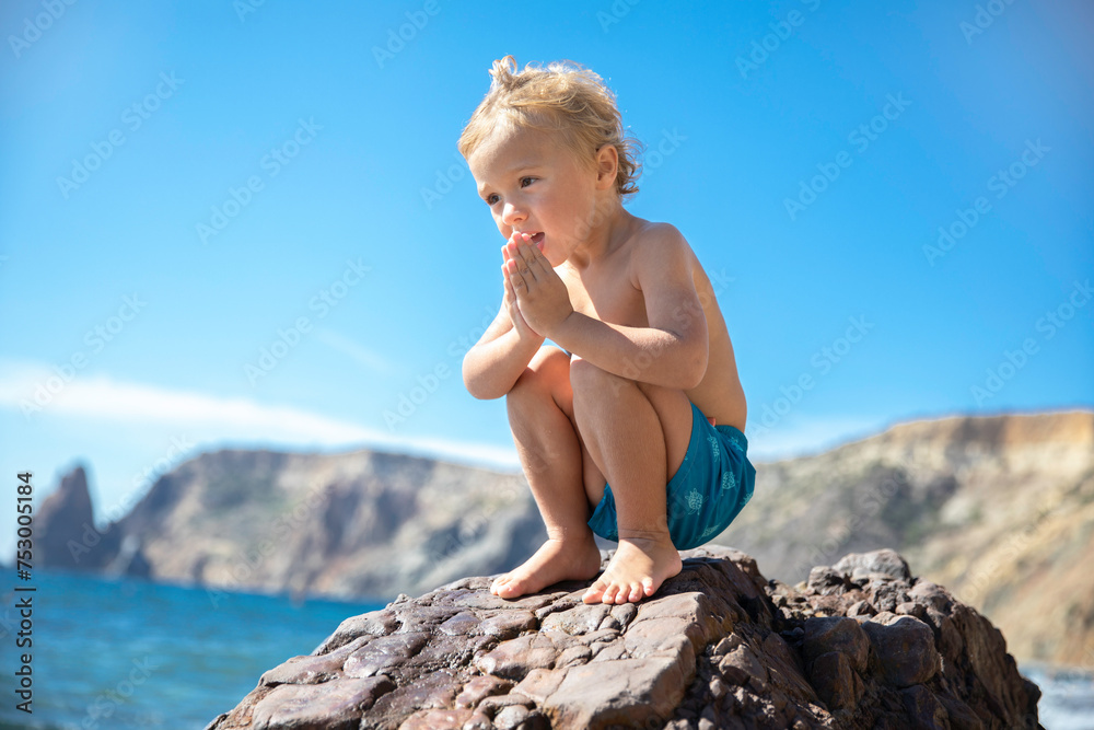 A small blond boy in swimming trunks plays on a stone on the seashore on a warm sunny day. A small blond boy in swimming trunks plays on a stone on the seashore on a warm sunny day.