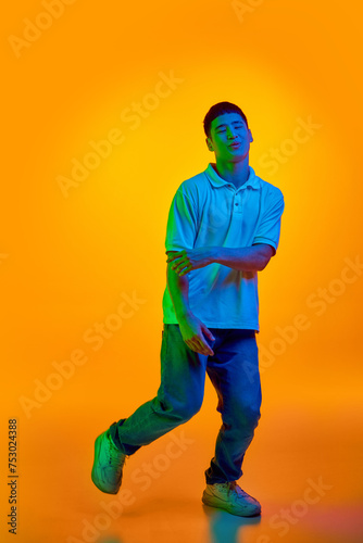 Full body photo of funny, cheerful, young Asian man dancing with joy in motion in vibrant neon light against gradient orange background. Concept of human emotions, fashion and beauty, work and hobby.