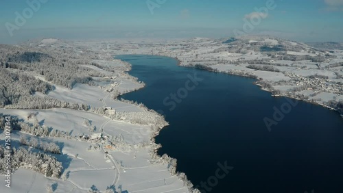 Aerial shot of a picturesque winter landscape over lake Irrsee in Austria during a sunny day, blue sky and snowy fields surrounding the lake.