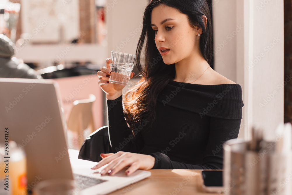 Smart caucasian woman with glass of water in hand studying with laptop. Student teen girl sitting in cafe drinks glass of water for body hydration while studying. Healthy lifestyle theme. Smart caucasian woman with glass of water in hand studying with laptop. Student teen girl sitting in cafe drinks glass of water for body hydration while studying. Healthy lifestyle theme.