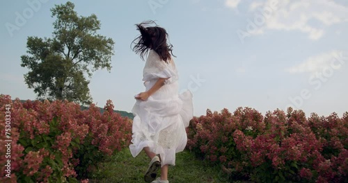 Back shot of woman running across green meadow full of blooming flowers.