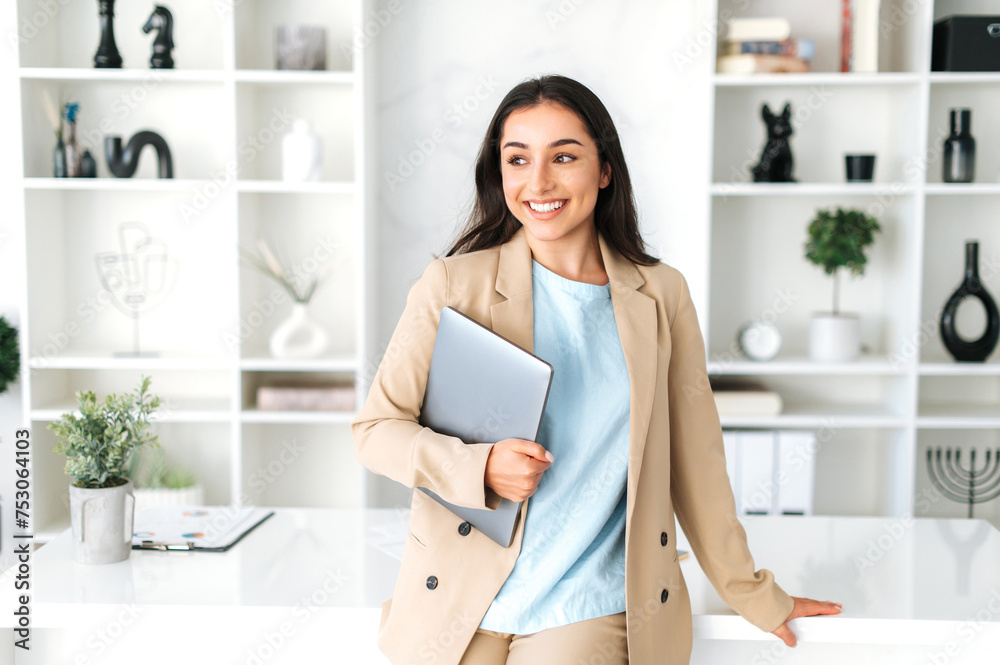 Successful cute arabian or indian young business woman in a stylish suit, digital manager, seo, holding laptop while standing in a modern office space near work desk, looking and smiling at camera Successful cute arabian or indian young business woman in a stylish suit, digital manager, seo, holding laptop while standing in a modern office space near work desk, looking and smiling at camera