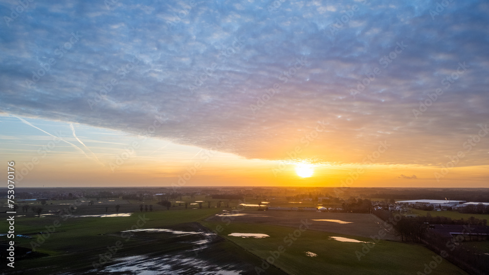 This aerial image captures the tranquil beauty of dawn as the sun breaks through a cloudy sky, casting a warm light over the wet fields below. The landscape is a patchwork of greens and dark This aerial image captures the tranquil beauty of dawn as the sun breaks through a cloudy sky, casting a warm light over the wet fields below. The landscape is a patchwork of greens and dark