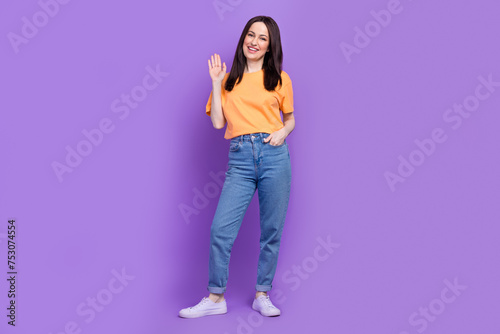 Full length body photo of sales manager woman in orange t shirt waving palm saying hi to customers isolated on purple color background