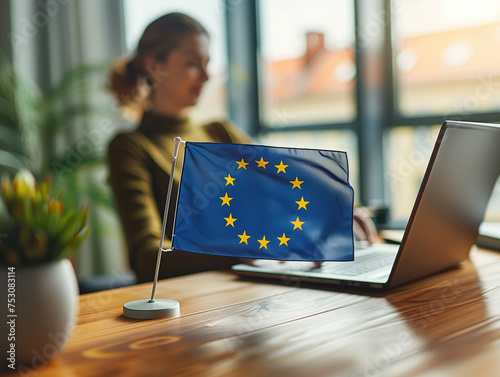 A European Union flag stands prominently on a desk with a focused individual working on a laptop in the background. © Александр Марченко