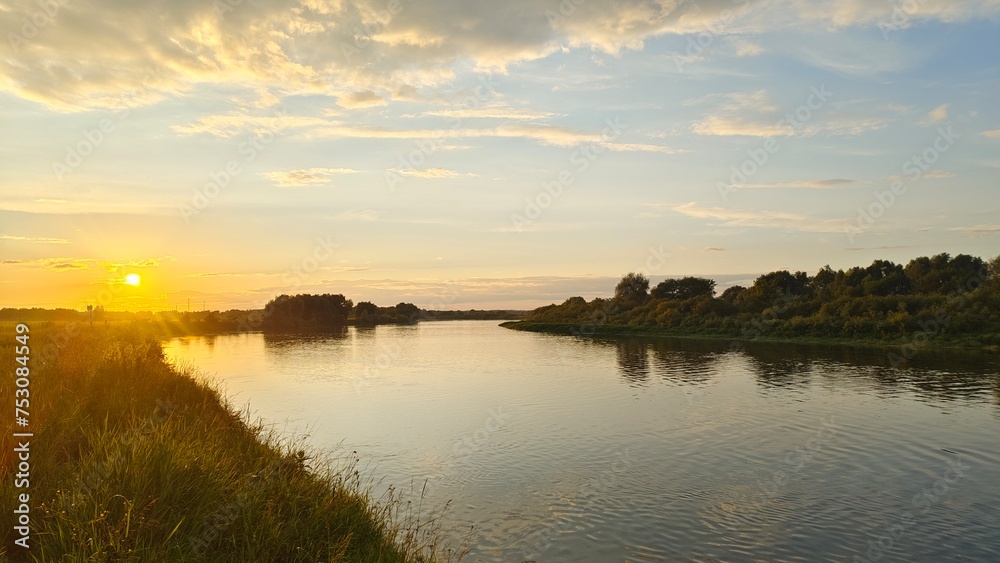 On a summer evening, the sun sets over the horizon and paints the sky in bright colors There is a ripple on the water Tall grass and deciduous trees grow along the banks and are reflected in the water