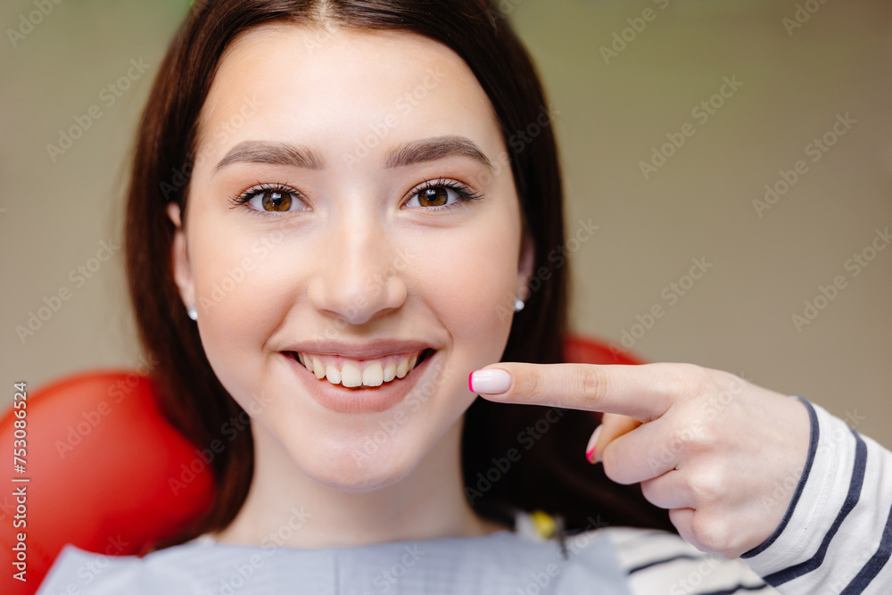 Cropped close up photo of young smiling girl, covered by napkin, pointing index finger on healthy toothy smile sitting at dental office chair. Healthcare teeth treatment Cropped close up photo of young smiling girl, covered by napkin, pointing index finger on healthy toothy smile sitting at dental office chair. Healthcare teeth treatment