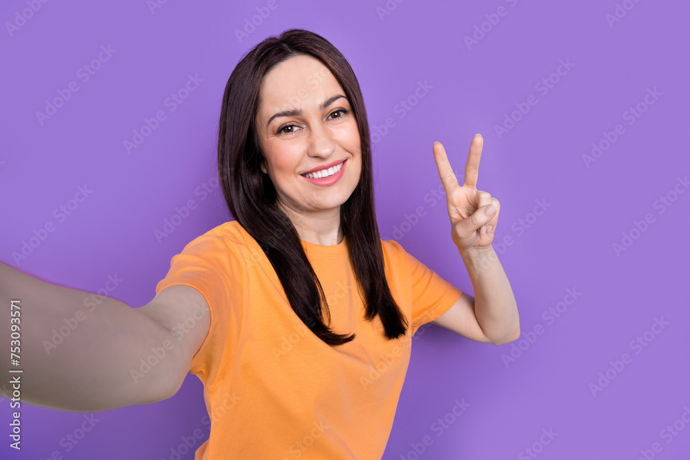 Selfie of smiling friendly woman in orange t shirt show v sign shooting typical vlog social media isolated on purple color background Selfie of smiling friendly woman in orange t shirt show v sign shooting typical vlog social media isolated on purple color background
