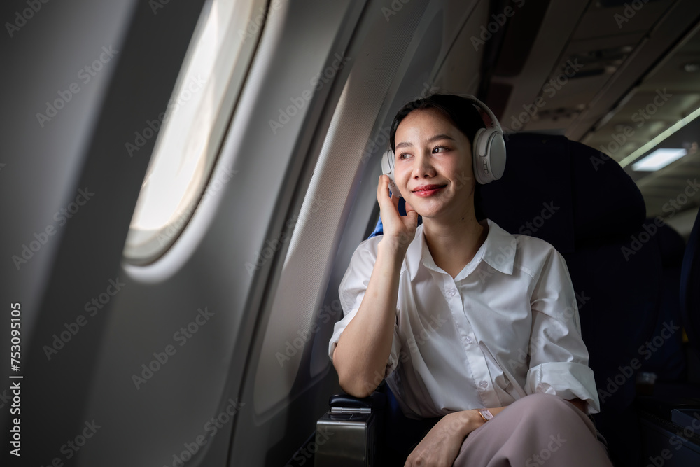 Young woman wearing headphones listening to music during travel, sitting near window in first class on airplane during flight, travel and business concept