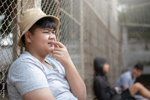 Asian teenboy in white t-shirt sits with picking teeth against a metal fence panel in a juvenile detention facility, awaiting further release, freedom and detention of people concept. photo