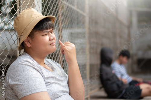 Asian teenboy in white t-shirt sits with picking mucus against a metal fence panel in a juvenile detention facility, awaiting further release, freedom and detention of people concept. photo