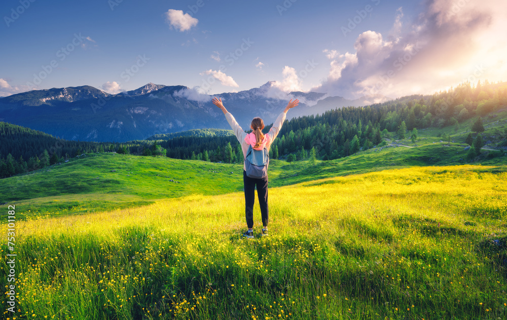 Happy girl on the hill with yellow flowers and green grass in beautiful alpine mountain valley at sunset in summer. Landscape with young woman with raised up arms in alps, trees, sky with clouds Happy girl on the hill with yellow flowers and green grass in beautiful alpine mountain valley at sunset in summer. Landscape with young woman with raised up arms in alps, trees, sky with clouds
