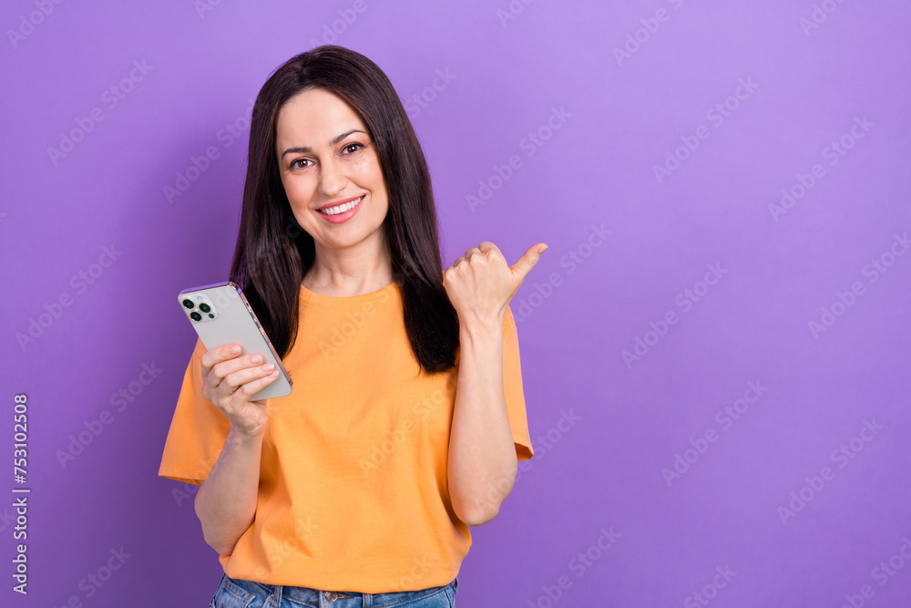 Photo of cheerful woman dressed orange t-shirt indicating at offer empty space hold smartphone isolated on purple color background Photo of cheerful woman dressed orange t-shirt indicating at offer empty space hold smartphone isolated on purple color background