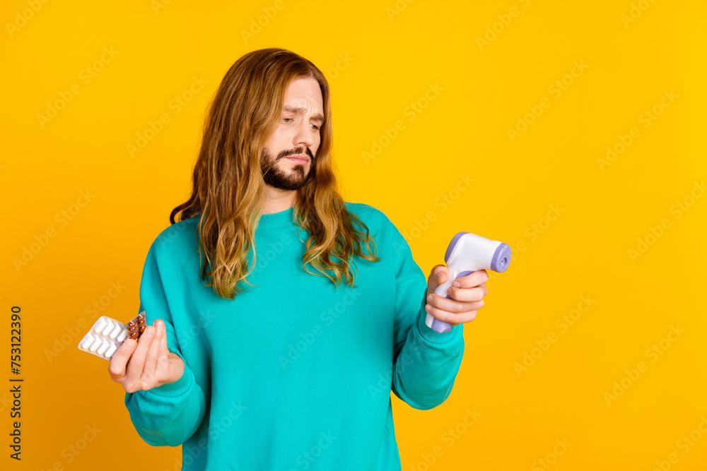 Photo of dissatisfied guy hold pills non contact thermometer empty space isolated on yellow color background Photo of dissatisfied guy hold pills non contact thermometer empty space isolated on yellow color background