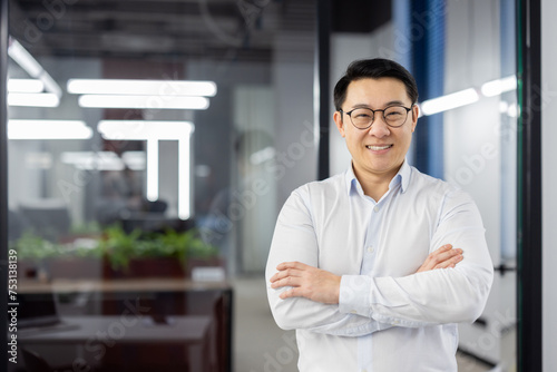 A professional Asian businessman stands confidently with arms crossed, smiling in a modern office setting.