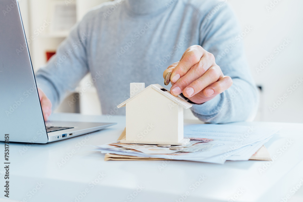 Close up male hand putting coin into House model money box on table with money and documents. Housing cost and payments. Real estate, property investment. Home mortgage loan rate. Saving money. Close up male hand putting coin into House model money box on table with money and documents. Housing cost and payments. Real estate, property investment. Home mortgage loan rate. Saving money.