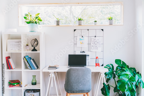 Modern cozy light workplace - white desk with laptop black screen, grid mood board with pinned notes, shelves with docs and green monstera plant at work space in home office room interior photo