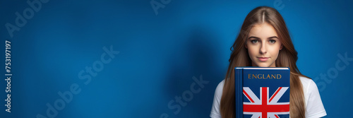 Cute schoolgirl holding blu English book on blue background. Copy space banner. Speak english and learn language concept
