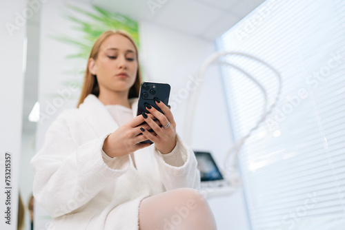Low-angle view of pretty blonde female patient in white uniform using typing smartphone in modern health center while waiting beautician specialist. Concept of cosmetology, spa, beauty treatment.