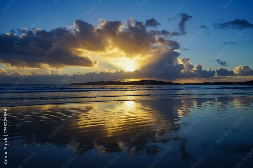 Les nuages se miroitent sur le sable humide à marée basse, peignant une toile céleste sur une plage de la Presqu'île de Crozon en Bretagne, offrant une vision saisissante de la nature.