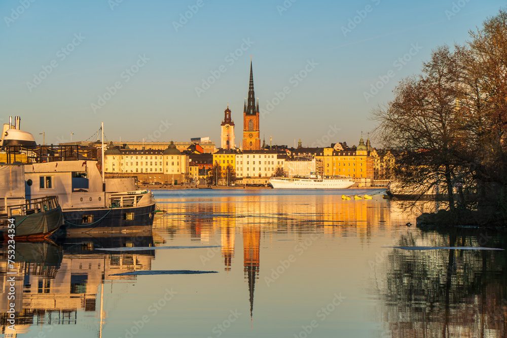 Fototapeta premium Riddarholmen church reflection in the lake at sunset. Stockholm, Sweden. Blue sky, boats in foreground, late winter. Yellow buildings.