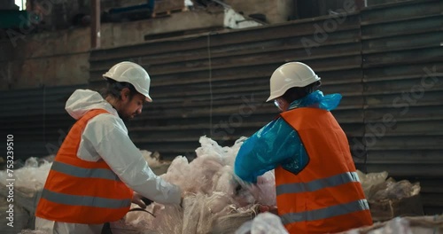 A brunette man with a beard in a white protective uniform and an orange vest together with a brunette girl presses plastic cellophane while working at a waste processing and sorting plant photo
