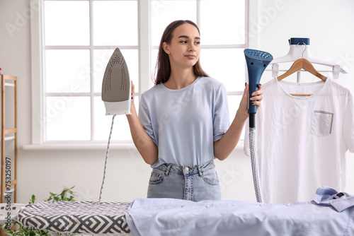 Canvas-taulu Pretty young woman with modern iron and garment steamer in laundry room