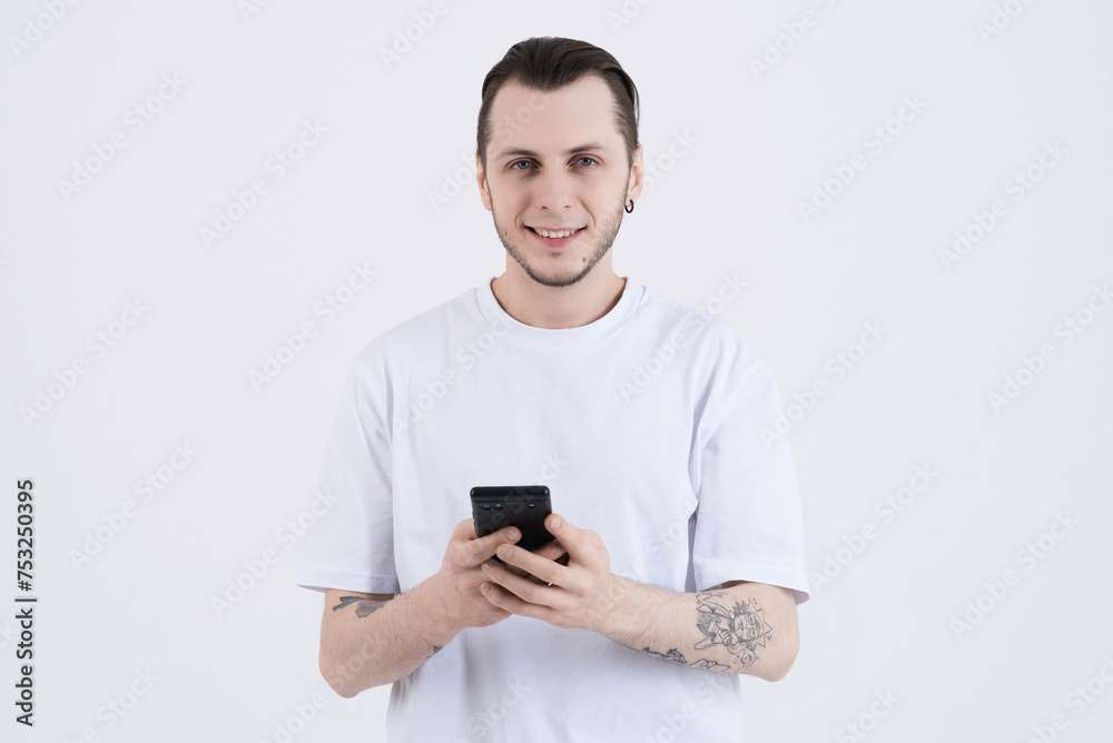 Attractive caucasian young man with tattoos in casual clothes confidently looking at camera and using mobile phone isolated on white studio background. Attractive caucasian young man with tattoos in casual clothes confidently looking at camera and using mobile phone isolated on white studio background.