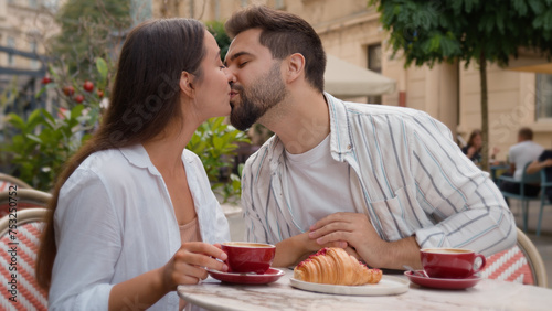 Young European couple eating breakfast together happy man woman smiling sweet kiss holding hands lovers romance city outside cafe restaurant honeymoon dating tenderness marriage bonding gen z urban 