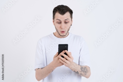 Wow! Attractive shocked caucasian young man with tattoos in casual clothes using mobile phone isolated on white studio background © Павел Костенко