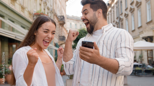 Caucasian couple happy woman man rejoices winning victory lucky expression together joy using mobile phone cellphone high-five exclamation success achievement emotions delighted triumph city outside