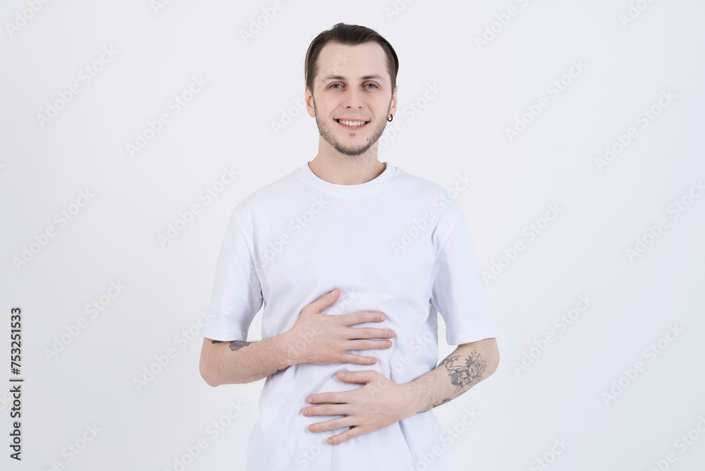 Attractive caucasian young man with tattoos in casual clothes smiling, looking at camera and with pleasure touching stomach isolated on white studio background Attractive caucasian young man with tattoos in casual clothes smiling, looking at camera and with pleasure touching stomach isolated on white studio background