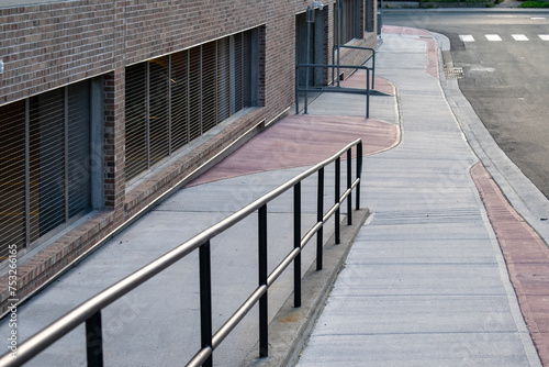 The exterior wall of a large brick building with a new concrete sidewalk. There's a wheelchair access ramp. There's a black metal handrail with a barrier free surface. The path is grey and pink color. photo