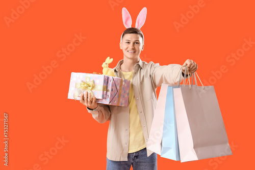 Young man in bunny ears with gifts and shopping bags on orange background. Easter Sale