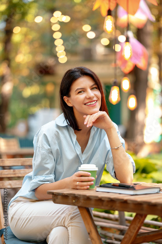 Young smiling woman sitting in park at cafe with a take away cup of coffee on a sunny day.