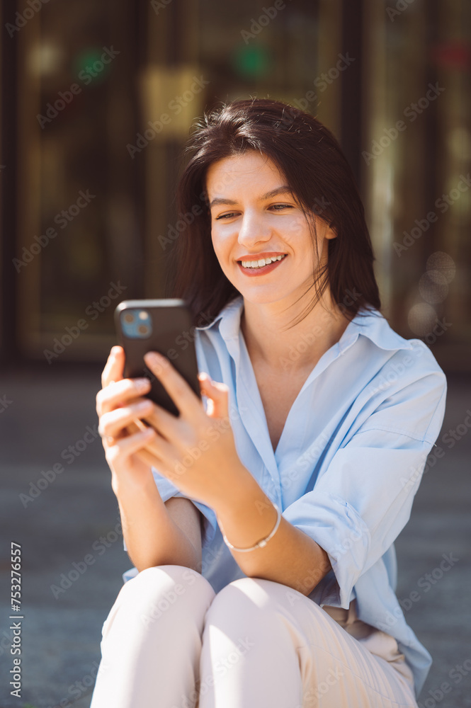 Wide smiling woman sitting on some stairs in the street is using her phone outdoors on a sunny day.