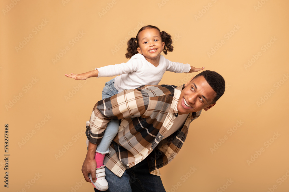 Funny african american young father playing with his little daughter, kid girl imitating airplane, spreading hands and smiling, beige studio background Funny african american young father playing with his little daughter, kid girl imitating airplane, spreading hands and smiling, beige studio background