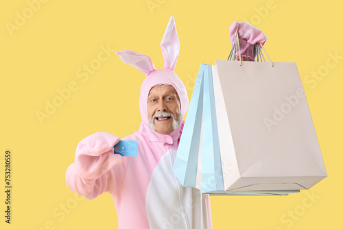 Senior man in Easter bunny costume with shopping bags and credit card on yellow background