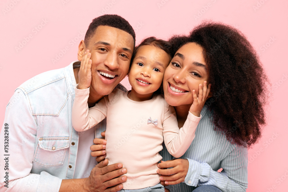 Happy black family of three with little daughter embracing, posing against pink studio background, parents and kid girl expressing love and togetherness Happy black family of three with little daughter embracing, posing against pink studio background, parents and kid girl expressing love and togetherness