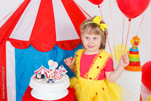 A little girl in a colorful outfit by a birthday cake. Circus theme, white, blue, red, yellow colors. Horizontal picture. 