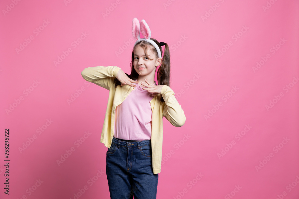 Adorable kid wearing bunny ears for easter photoshoot in studio, celebrating spring holiday over pink background. Cute little girl being playful and festive for traditional celebration.
