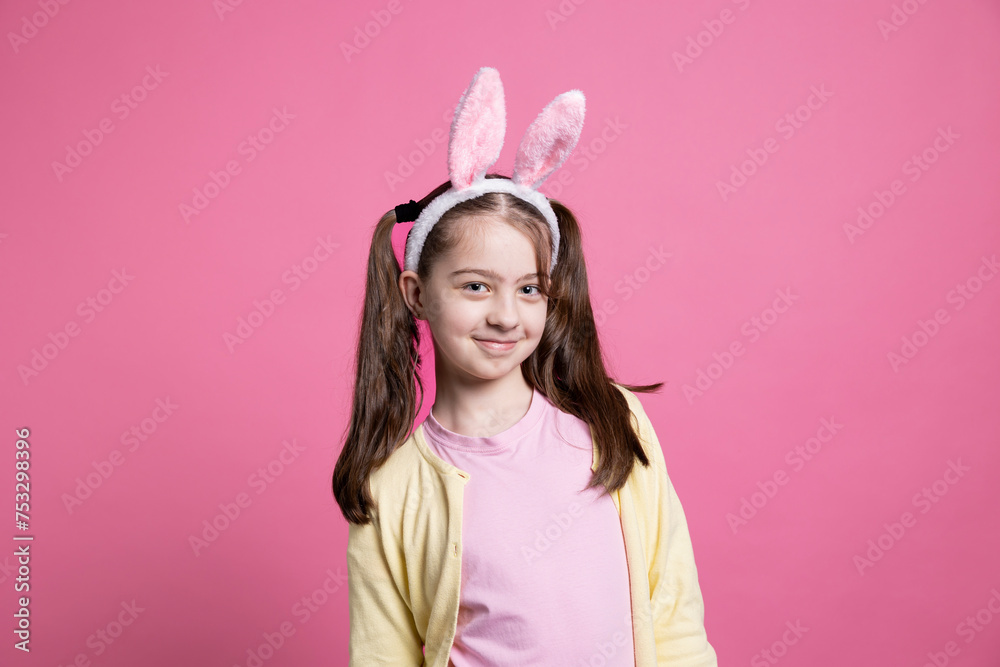 Young confident girl with pigtails and bunny ears posing on camera, feeling happy and joyful about easter celebration. Little kid being adorable over pink background, festive spring holiday.