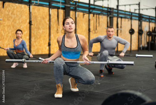 Fit young woman doing barbell squats exercise near other people at crossfit gym