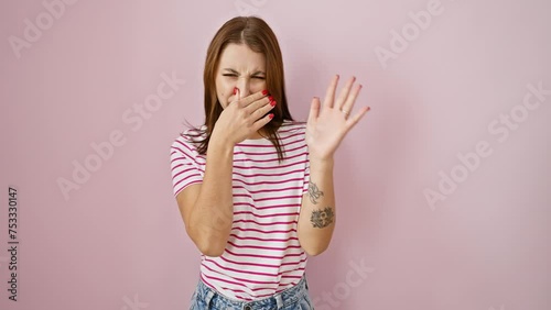 Young brunette girl in stripes t-shirt, holding breath over bad stink, disgust on nose from intolerable, stinky smell, isolated over pink background
