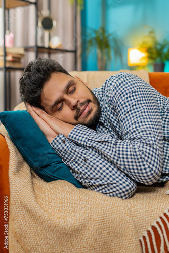 Tired Indian young man lying down in bed taking a rest. Carefree peaceful guy napping, falling asleep on comfortable sofa with pillows at home room. Closes his eyes enjoy daytime nap alone. Vertical