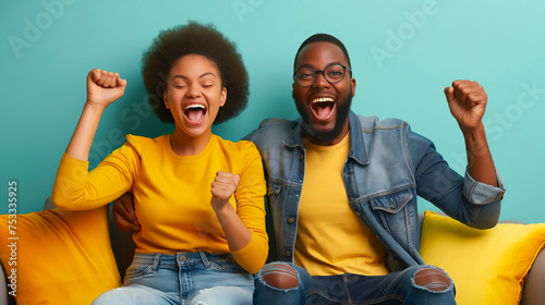 Excited African American couple celebrating success while sitting on sofa at home. Overjoyed happy young man and woman in casual wear raising clenched fists in excitement doing winner gesture photo