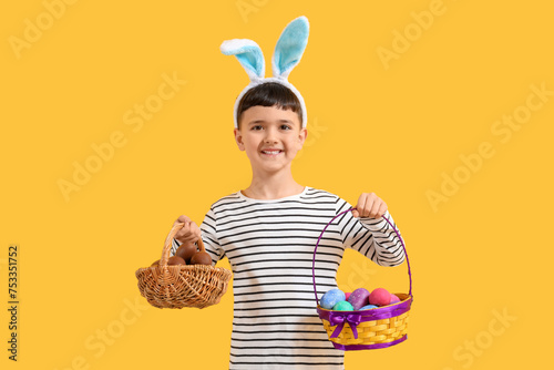 Cute little boy in bunny ears headband holding baskets with Easter egg on yellow background