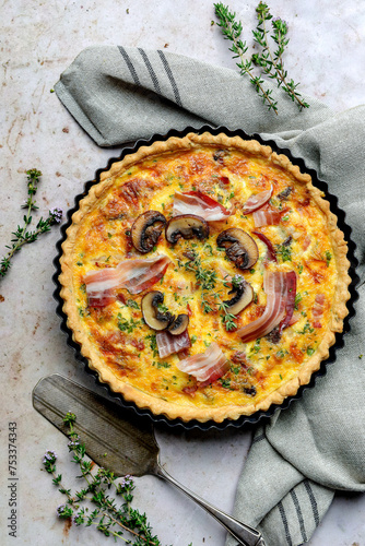 Traditional French quiche with prosciutto and mushrooms, captured in a rustic tin in an overhead portrait shot. Presented on a light background with grey teatowel and pie server photo