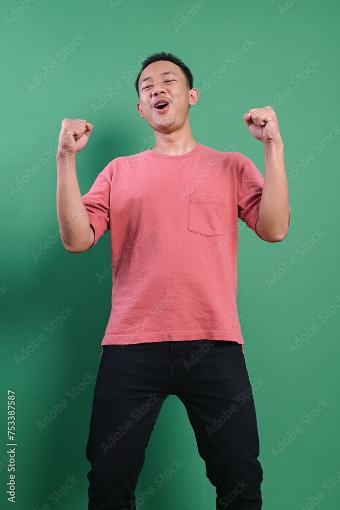 Cheerful Asian man celebrating victory raising both hands over green background Cheerful Asian man celebrating victory raising both hands over green background